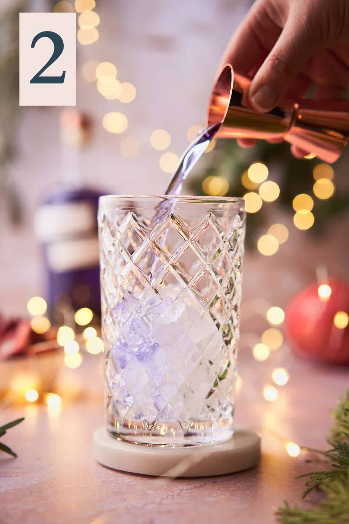 hand pouring purple gin into an ice filled cocktail shaker, with twinkling lights, pomegranate, and balsam in the background.