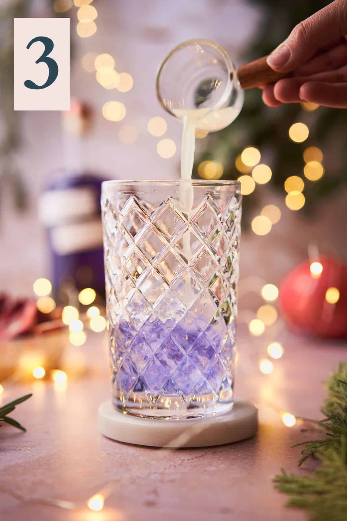 hand pouring lime juice into an ice filled cocktail shaker with purple gin, with twinkling lights, pomegranate, and balsam in the background.