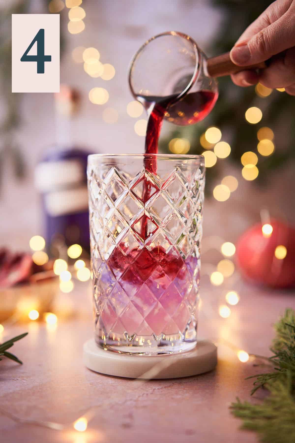 hand pouring pomegranate juice into an ice filled cocktail shaker with purple gin, with twinkling lights, pomegranate, and balsam in the background.