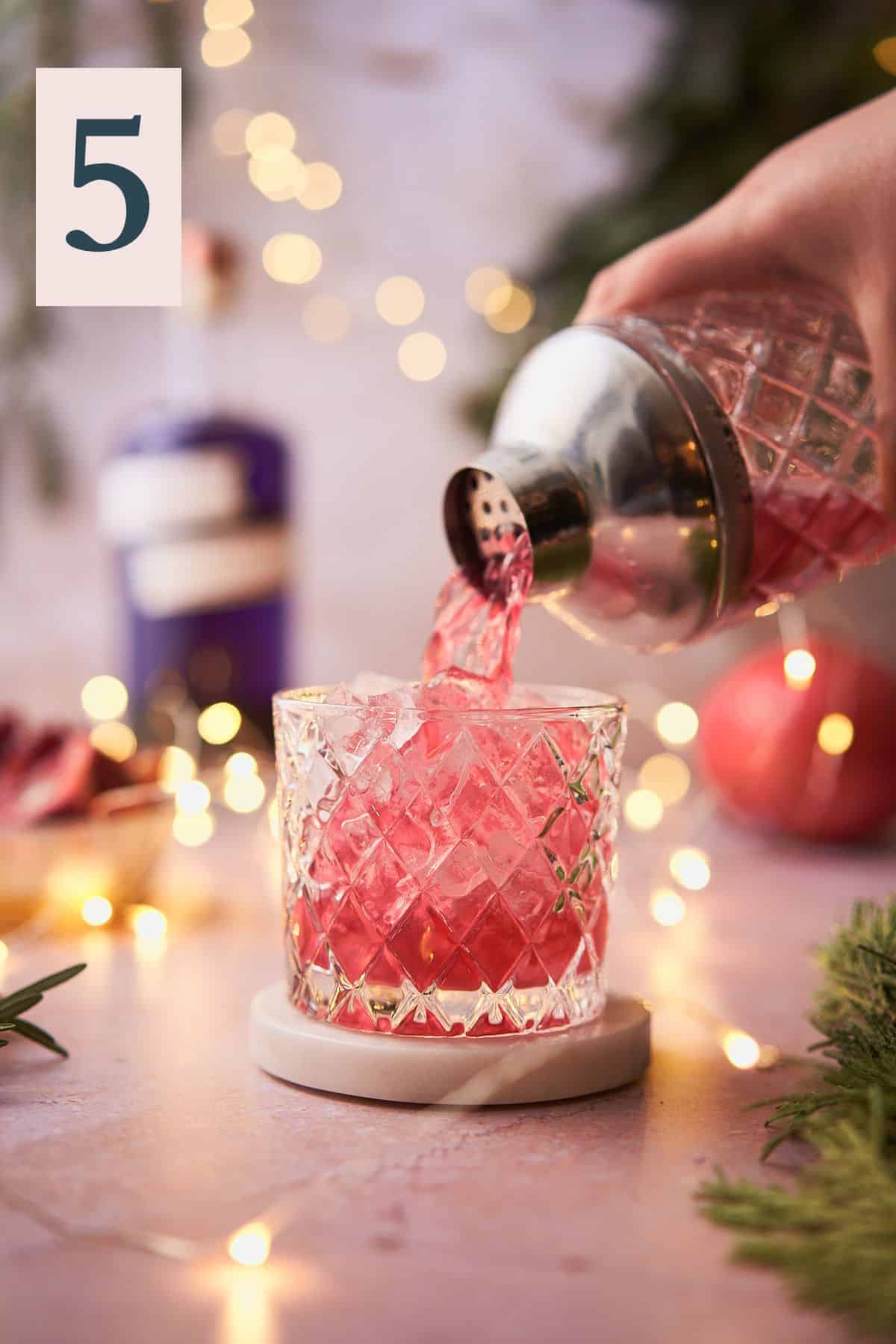 hand pouring a bright red cocktail mixture into an ice filled low ball glass, with twinkling lights, pomegranate, and balsam in the background.