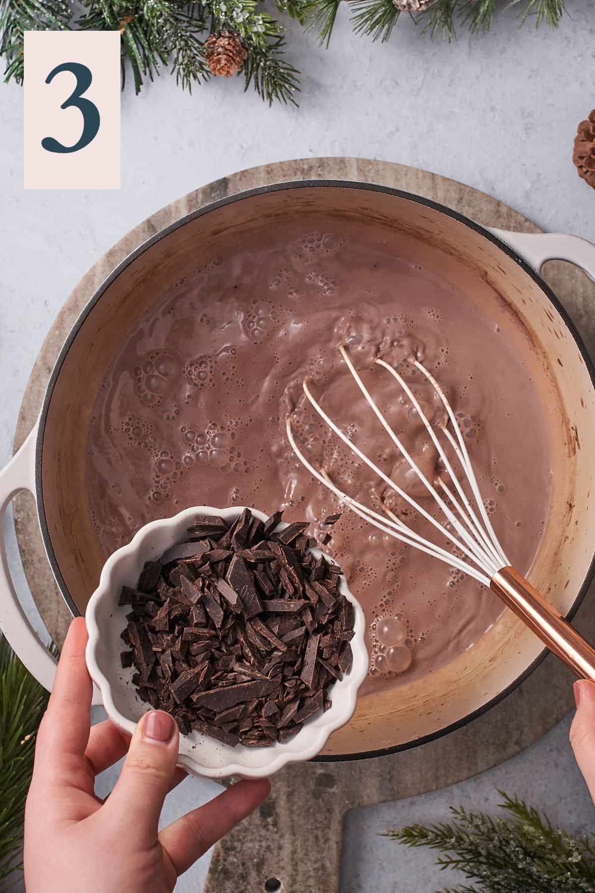 hand pouring chopped chocolate pieces into a dutch oven with hot chocolate inside. 
