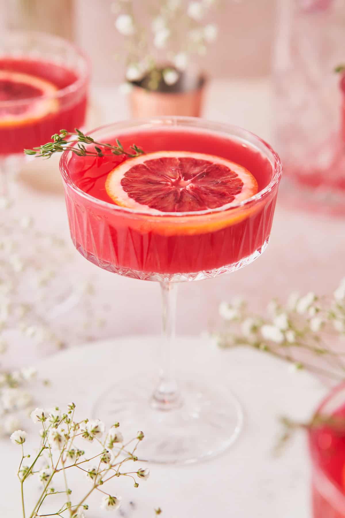 close up shot of a blood orange gin cocktail, surrounded by soft white baby's breath flowers, and other drinks in coupe glasses.