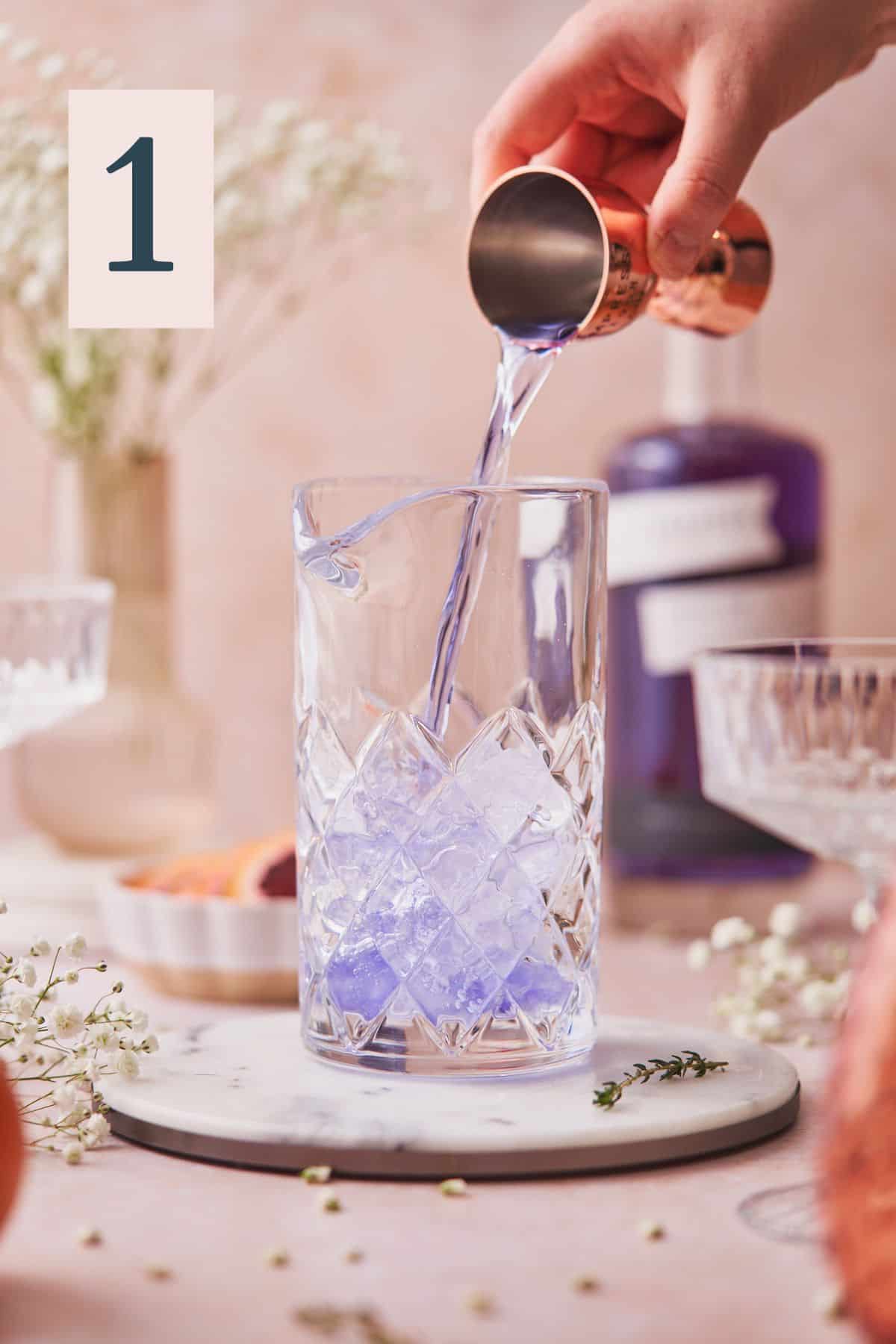 Pouring purple empress gin into a mixing glass with ice, and white flowers, blood oranges, and coupe glasses on the scene. 
