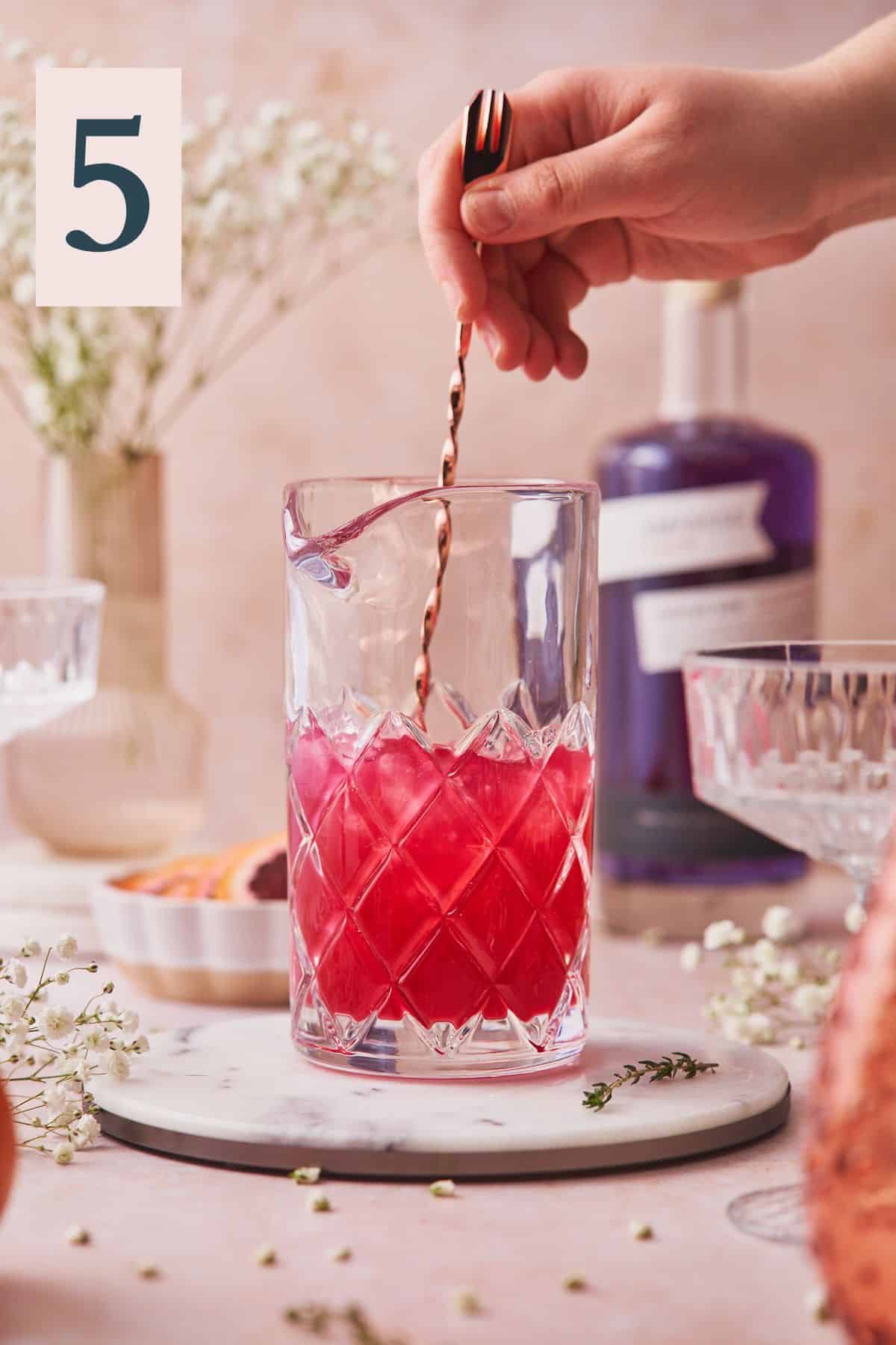 hand mixing a bright pink cocktail in a mixing glass surrounded by white flowers and cocktail glasses. 