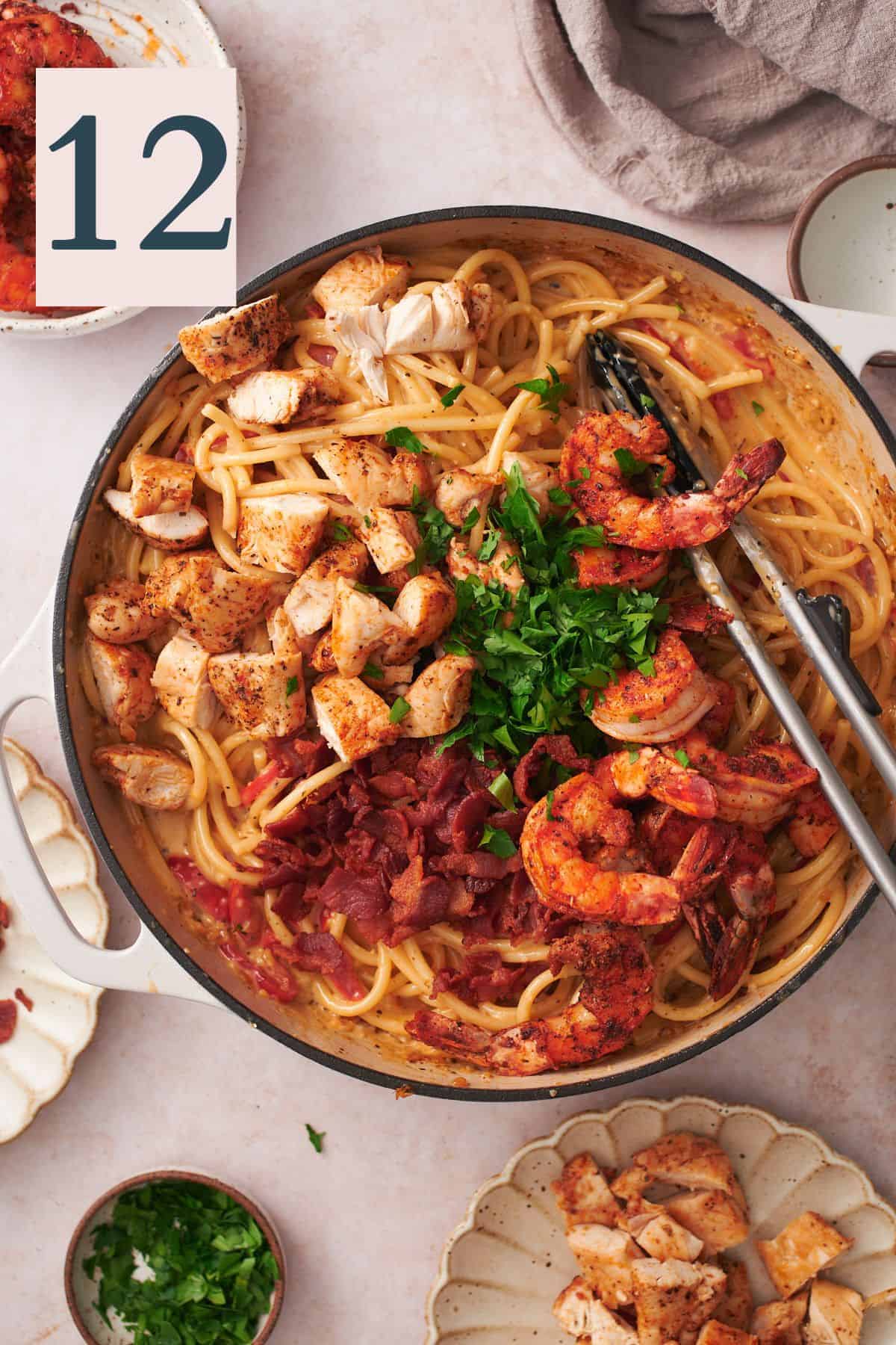 parsley, shrimp, bacon, and chicken added to an enameled skillet with pasta and tongs in the skillet. 