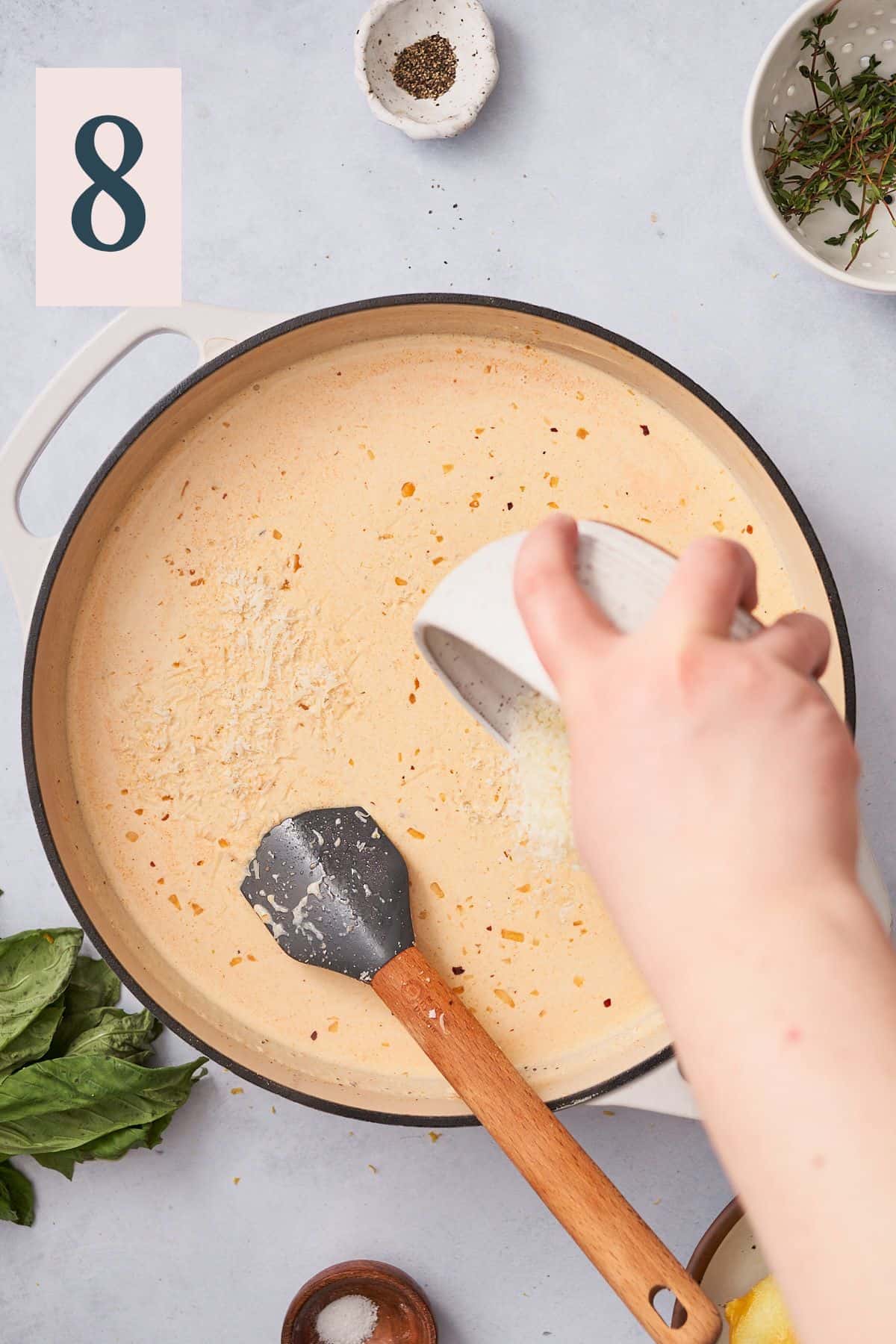 hand pouring parmesan cheese into a cream sauce in an enameled cast iron skillet.