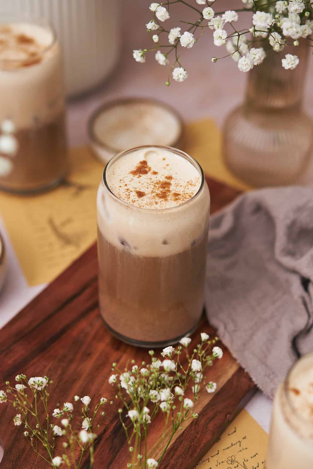 protein coffee in a glass jar with a frothy milk and pinch of ground cinnamon, with white baby's breath flowers in the foreground. 