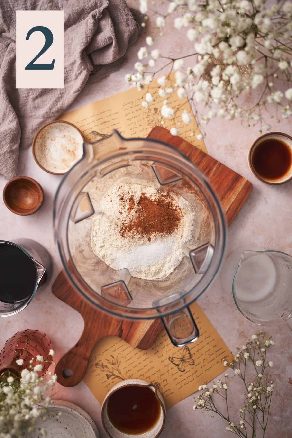 milk, protein powder, salt, and cinnamon in a blender jug, surrounded by other ingredients to make the drink, and white flowers.  