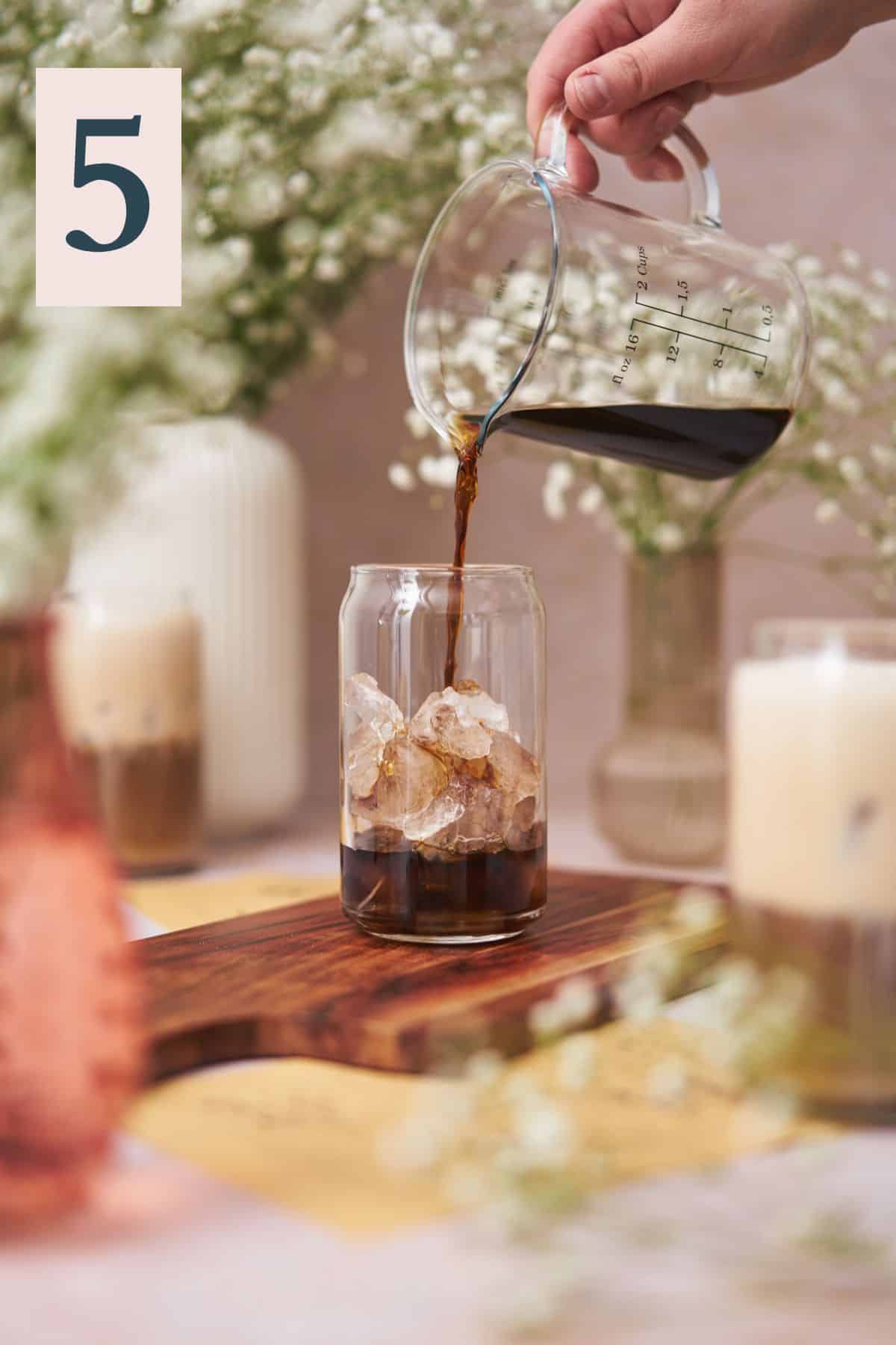 pouring iced coffee into a glass with ice, surrounded by glasses, and vases with small white baby's breath flowers.