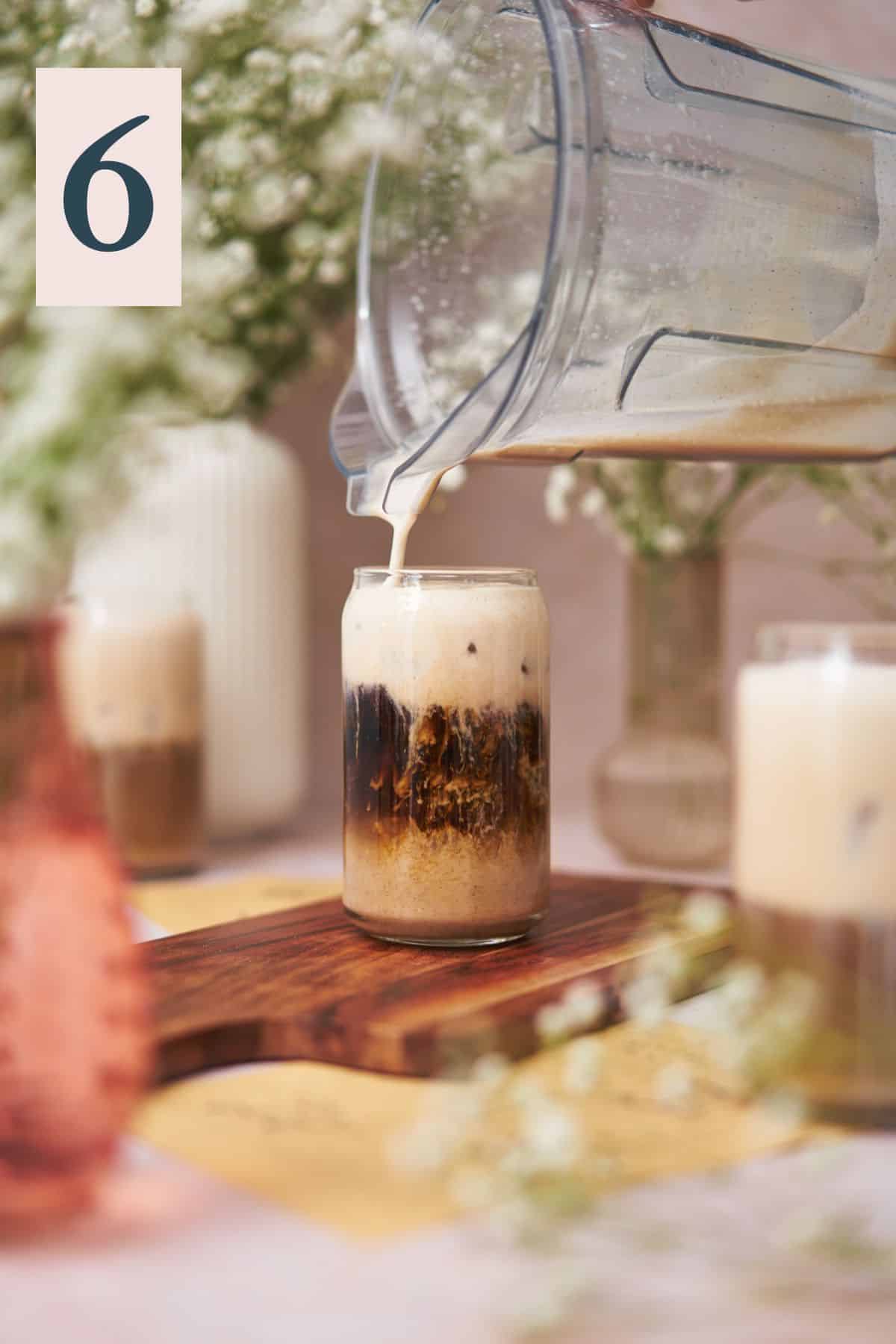 pouring a creamy white mixture into a glass with ice and coffee, surrounded by glasses, and vases with small white baby's breath flowers.