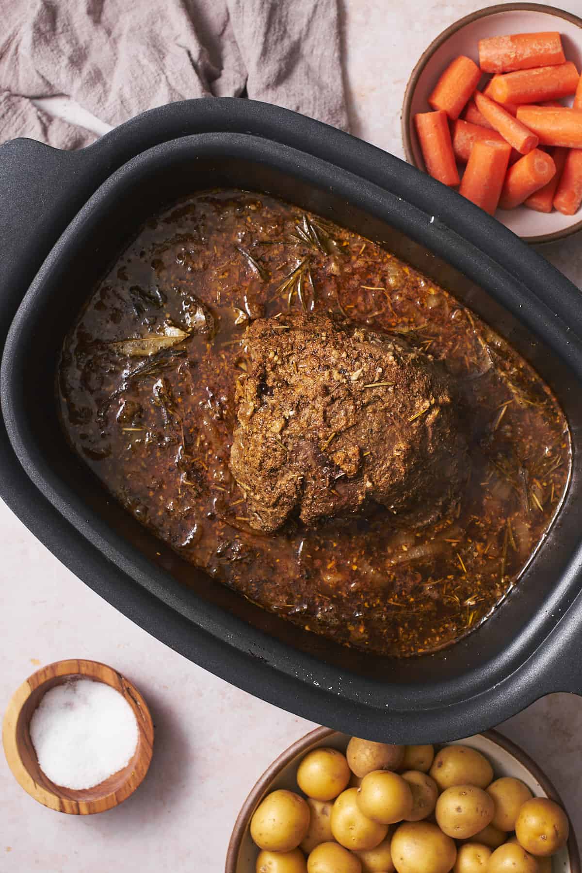 Partially cooked roast in a slow cooker with braising liquid and herbs.