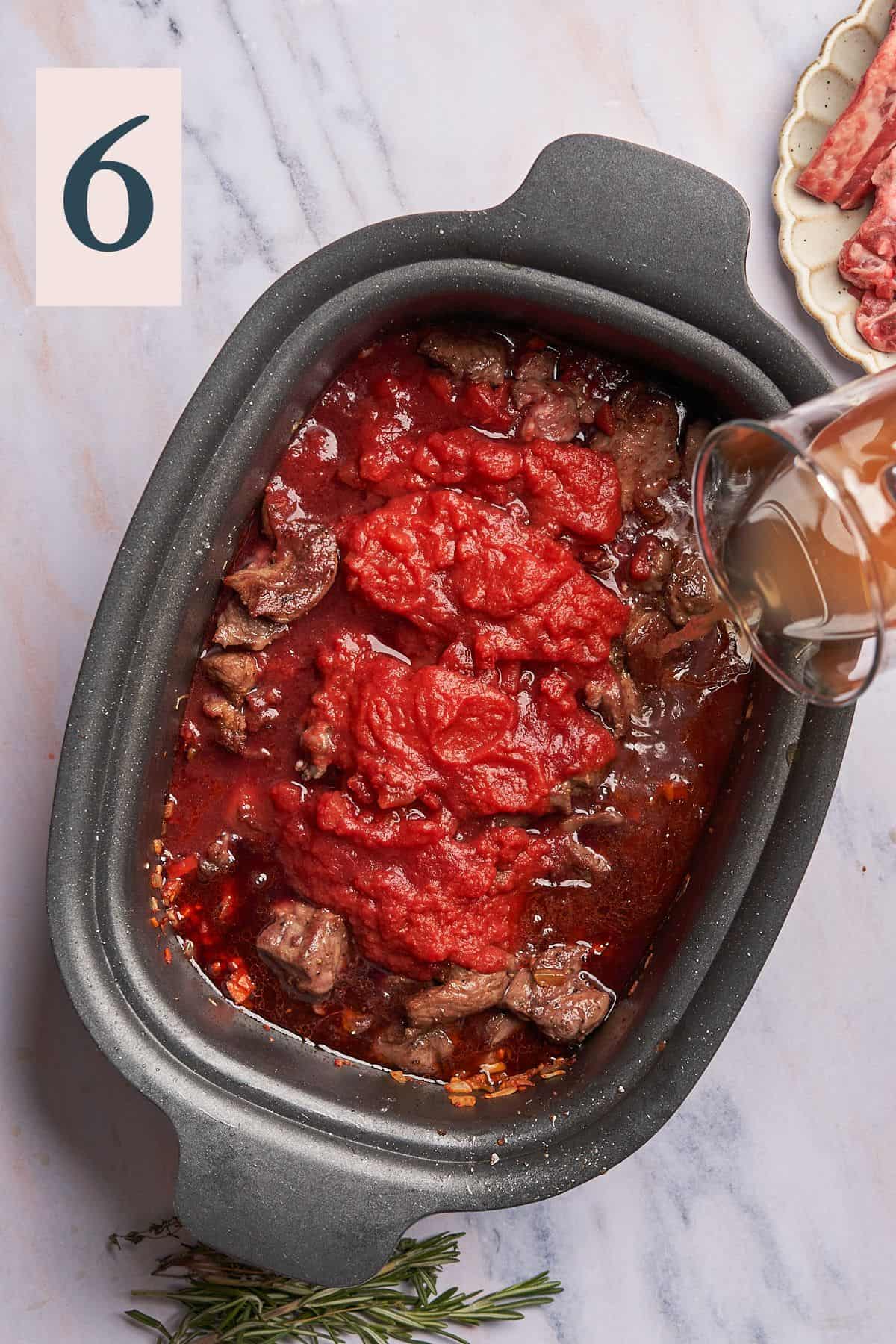hand pouring beef broth into a slow cooker with lamb, crushed tomatoes, and cooked vegetables. 