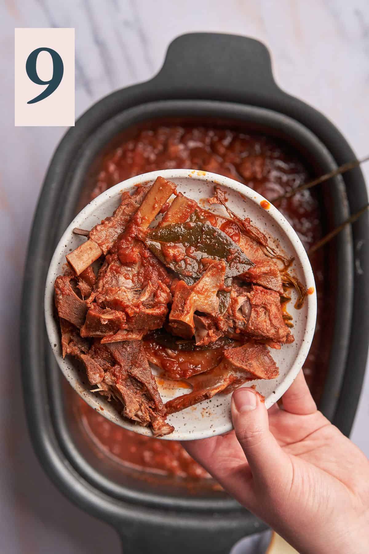hand holding a plate of lamb bones and bay leaves over a slow cooker of cooked lamb ragu. 