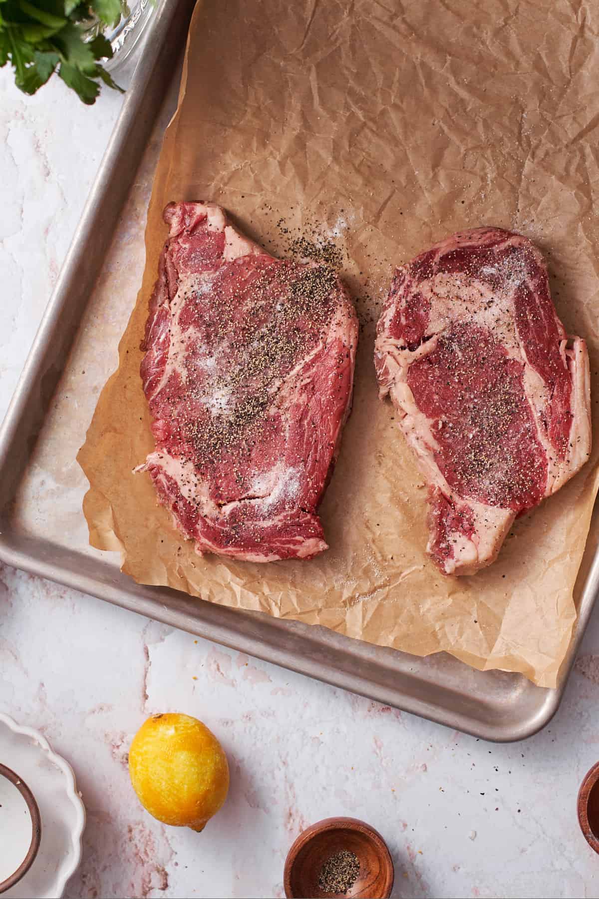 Ribeye steaks seasoned with salt and pepper on a parchment lined baking sheet.