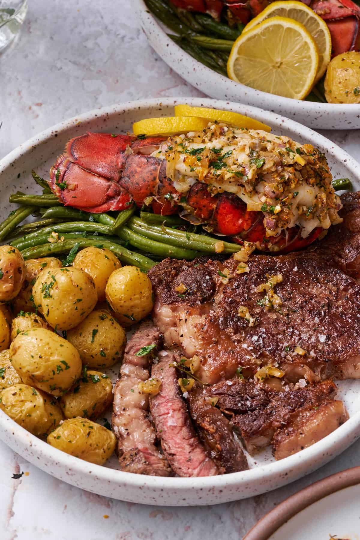 45 degree shot of a plate with a medium rare ribeye, green beans, roasted baby potatoes, and a cooked lobster tail with herbs and butter.