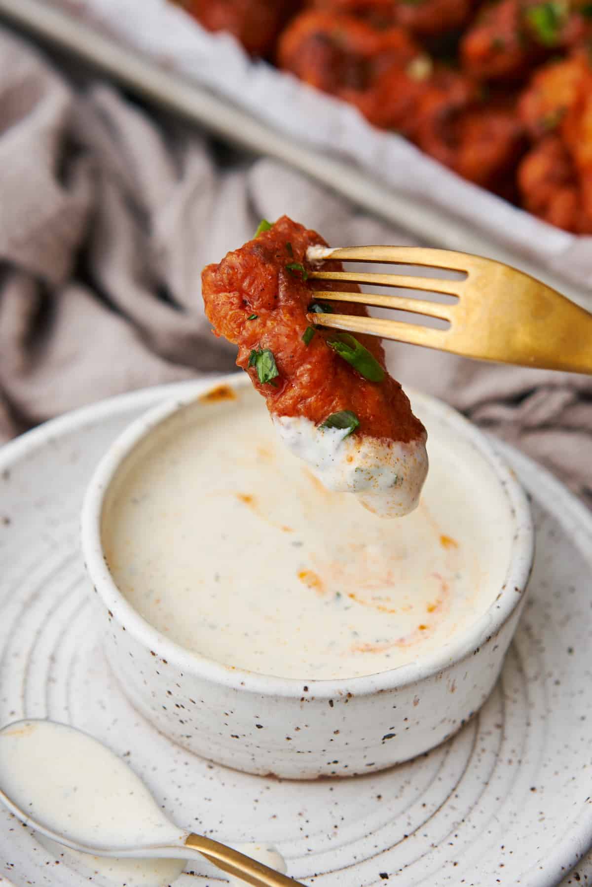 Boneless chicken wings being dipped in ranch dressing with a gold fork, and a tray of chicken wings in the background. 
