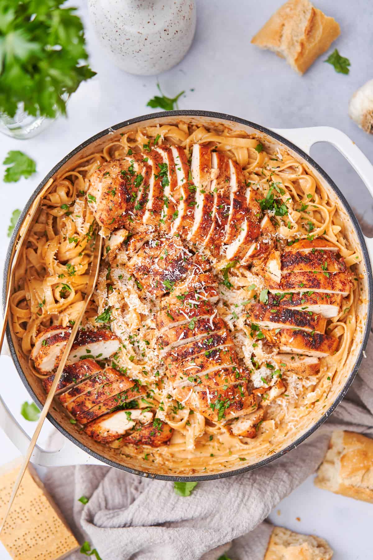 large enameled cast iron skillet with creamy cajun chicken alfredo and fettuccine in the pot, surrounded by parmesan, parsley, garlic, and a baguette. 