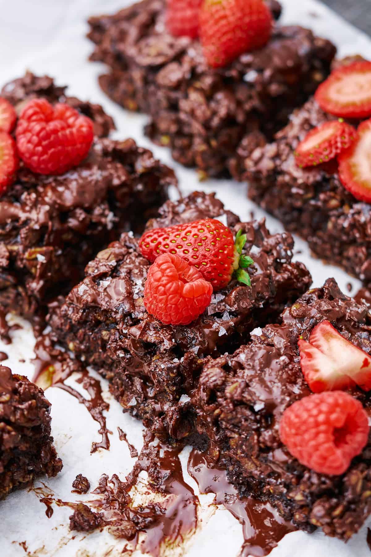 close up shot of chocolate baked oat bars topped with a chocolate drizzle, sea salt, and bight and juicy strawberries and raspberries. 