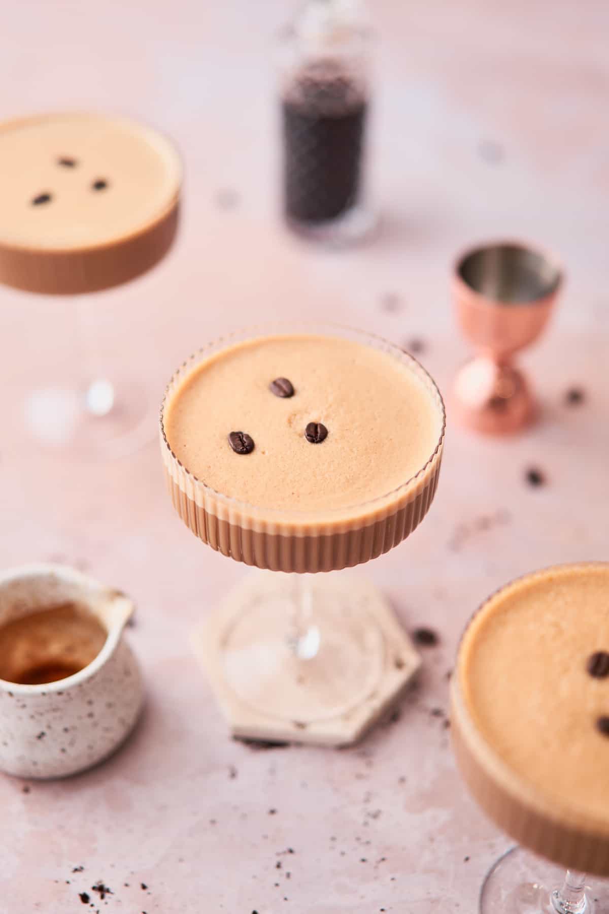 3 coupe glasses with creamy espresso martinis surrounded by coffee beans and various liqueurs. 