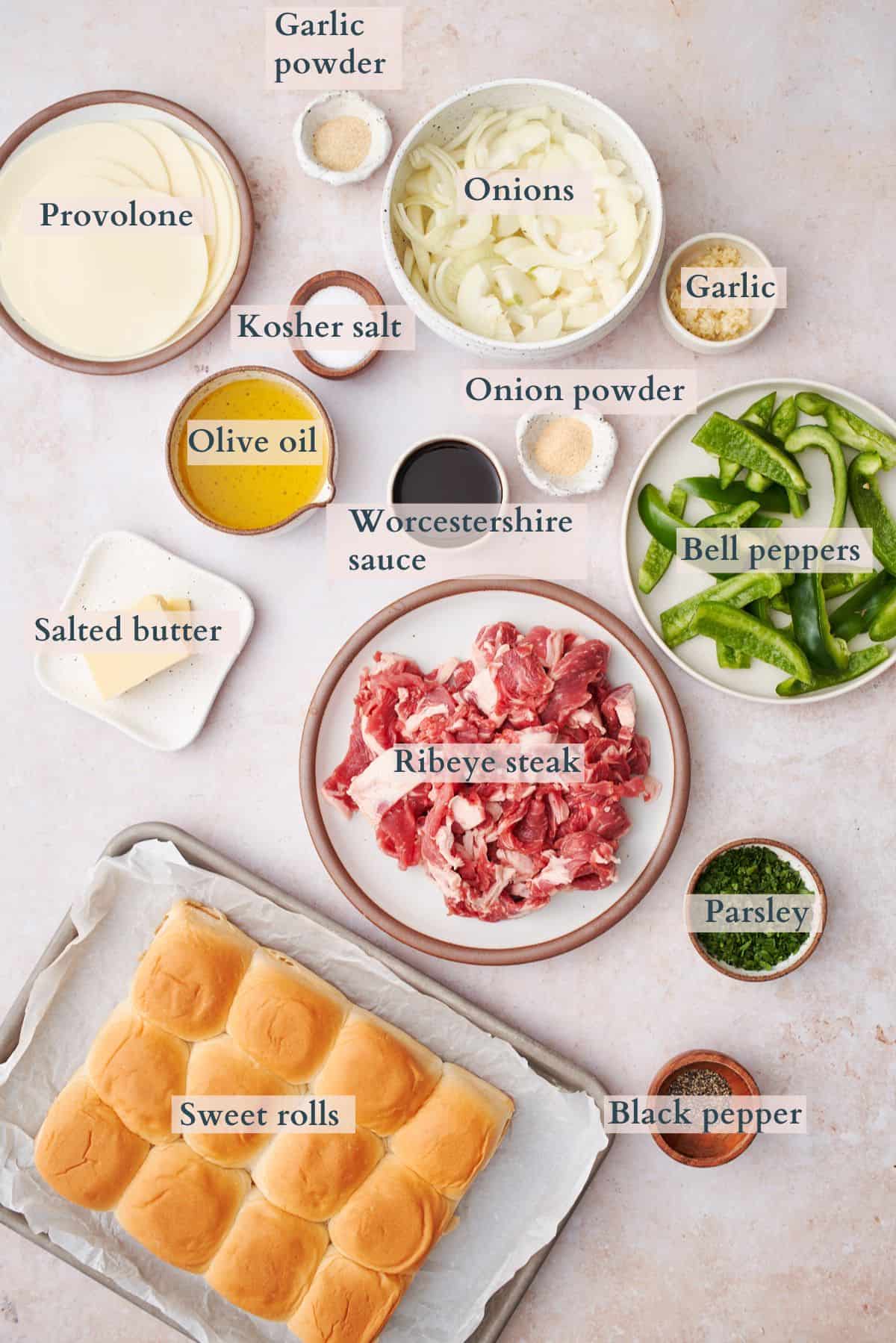 ingredients for cheese steak sliders laid on various plates and bowls.