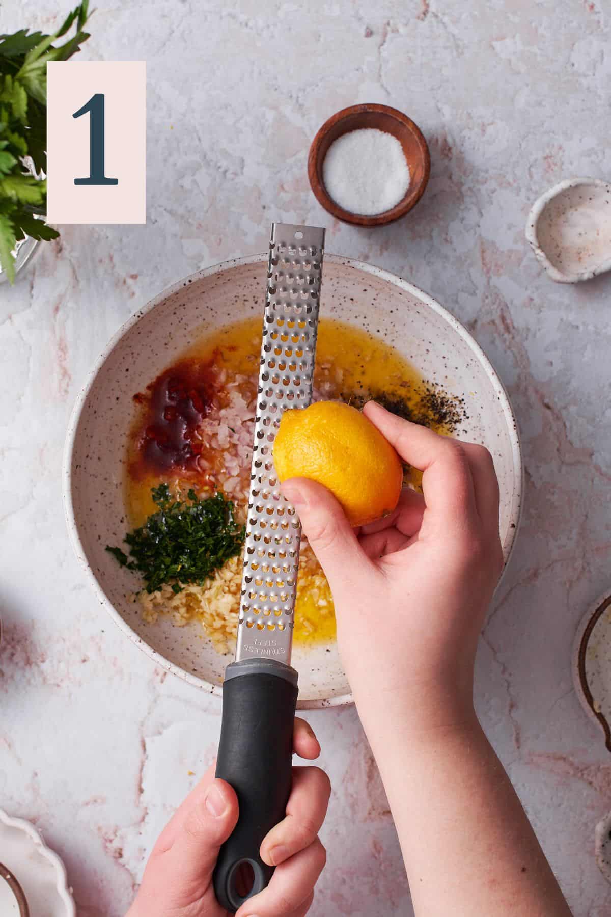 hand zesting lemon into a bowl with garlic, shallots, parsley, and spices in a bowl. 