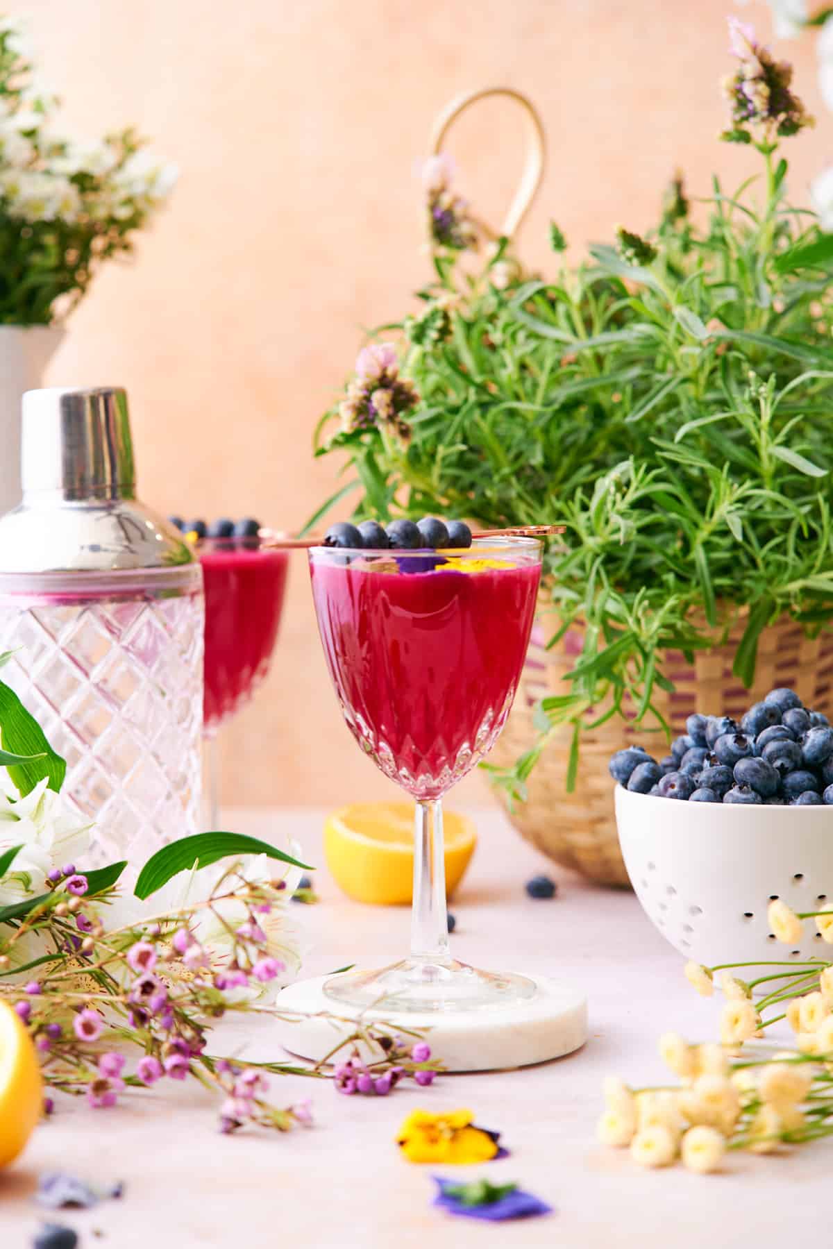 stunning straight on shot of a blueberry gin cocktail in a garden like scene, surrounded by fresh blueberries, lemons, and an assortment of flowers. 