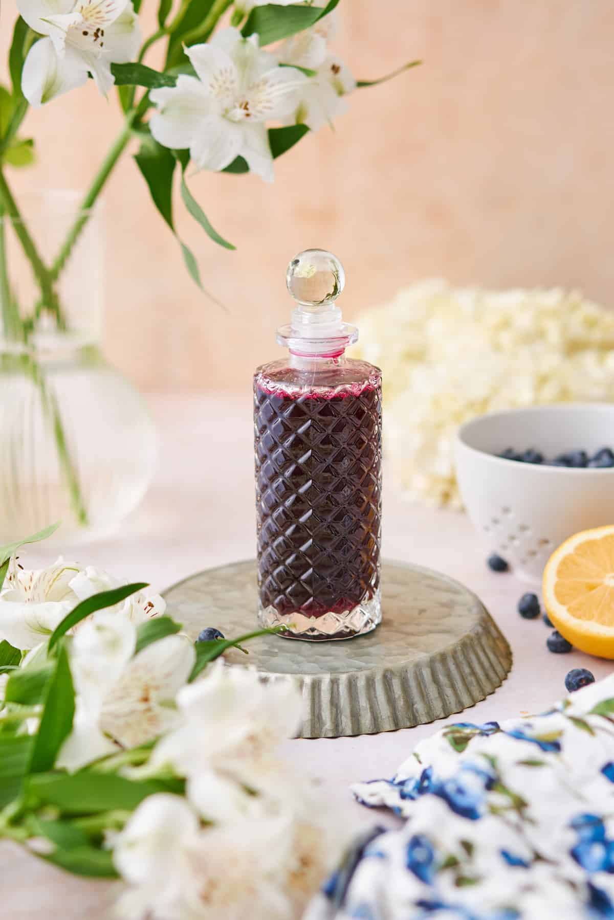 blueberry simple syrup in tall jar, surrounded by fresh berries, lemon, and white flowers. 