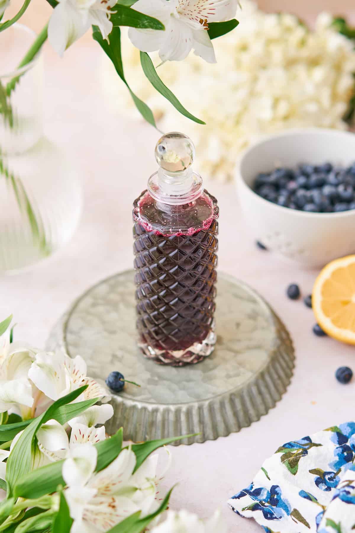 blueberry simple syrup in a tall, intricate glass container, with white flowers surrounding it, and fresh blueberries and lemon nearby. 