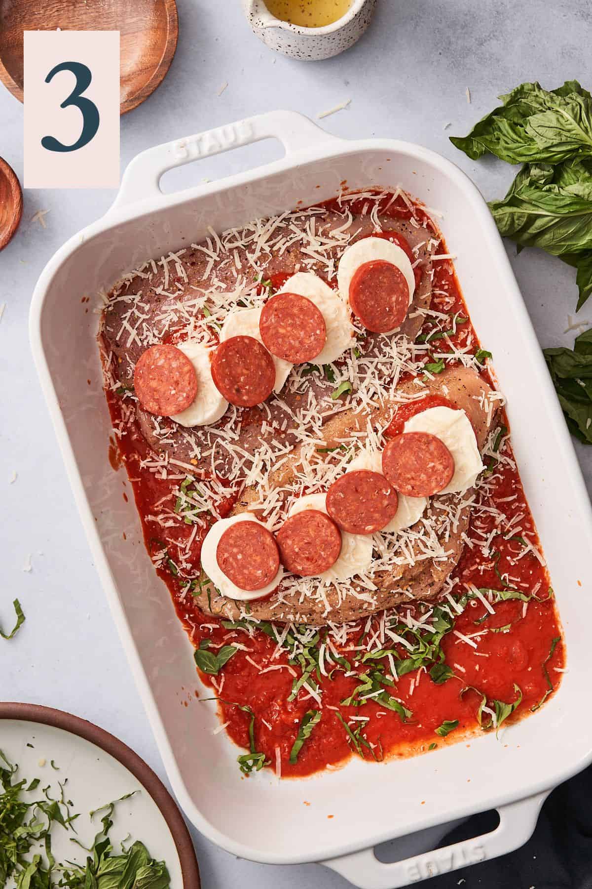 Casserole dish with open chicken breasts with sauce, cheese slices, parmesan, basil, and pepperoni inside of them.