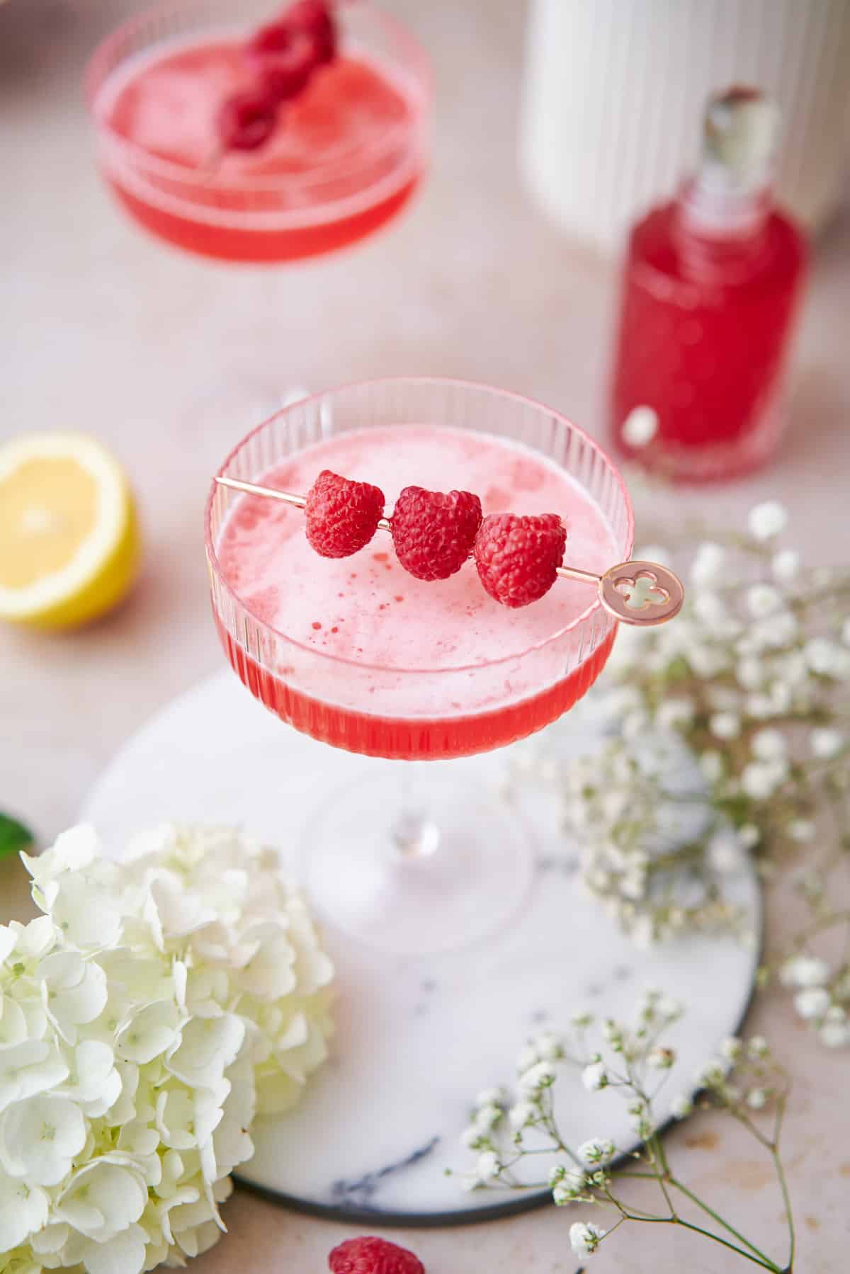 close up shot of a frothy raspberry martini surrounded by baby's breath flowers, lemon, and hydrangeas. 