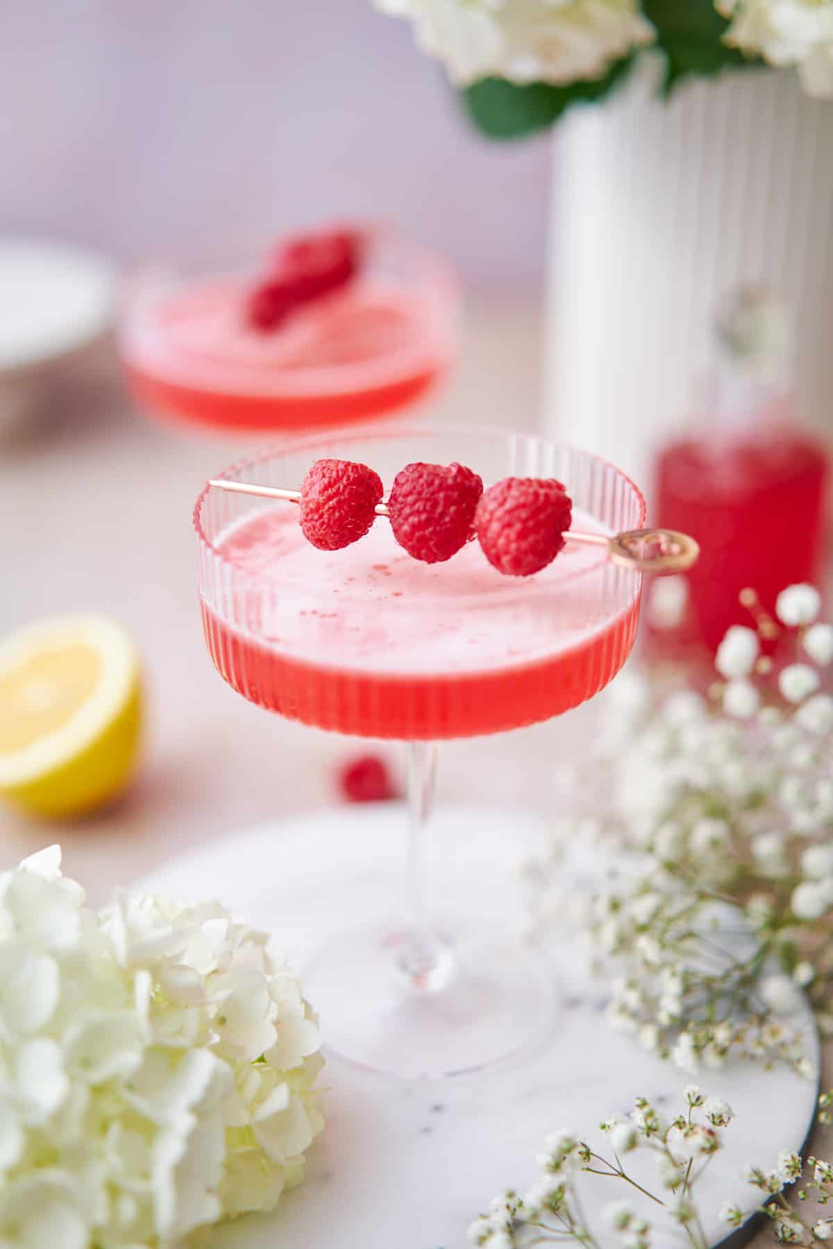 stunning raspberry martini in couple glass, with fresh raspberries, in a whimsical setting, surrounded by baby's breath flowers, and fresh lemons.