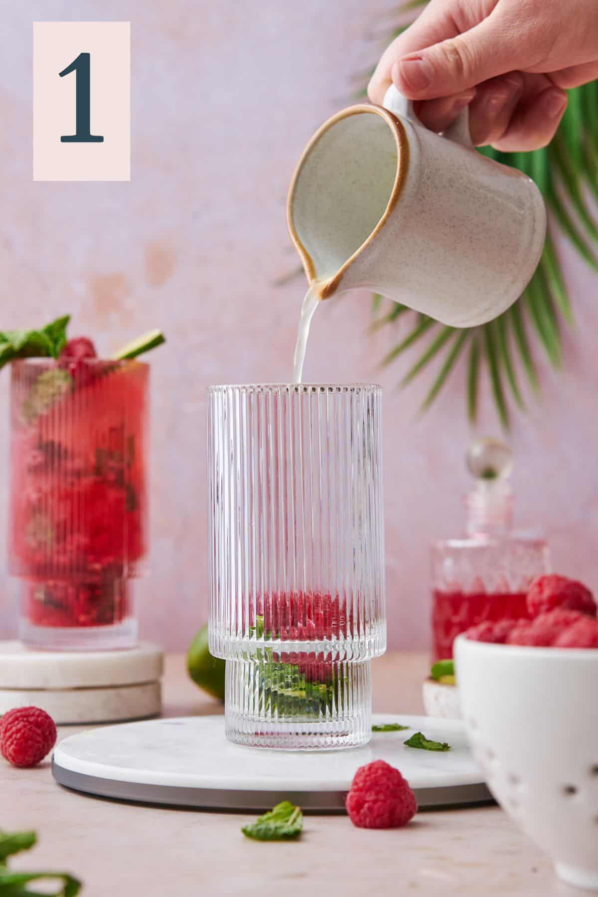 Pouring lime juice into a glass with raspberry and mint at the bottom.