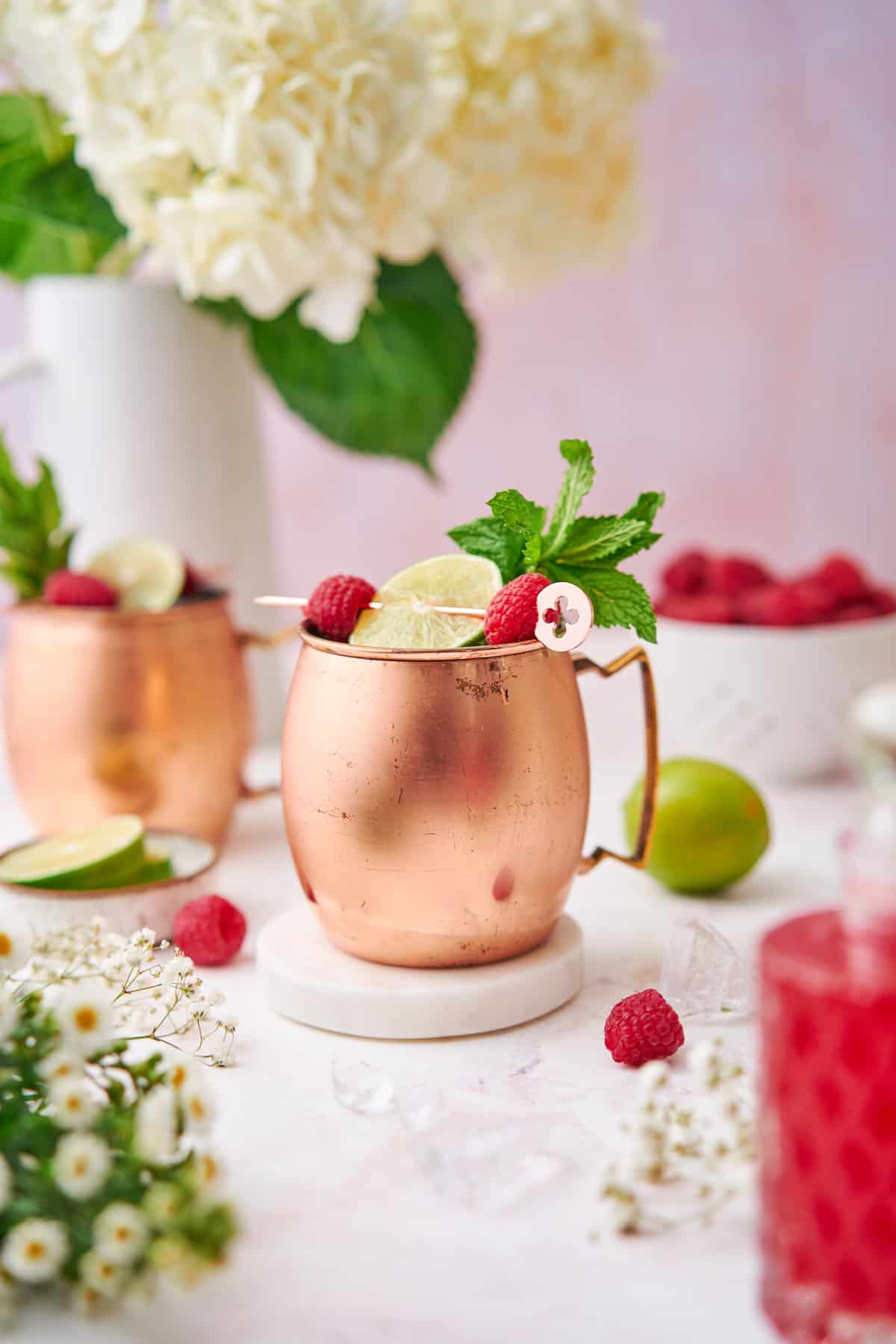 two copper moscow mule mugs with raspberry mules in the cups with fresh mint and lime wedges to garnish, surrounded by small white flowers, raspberries, limes, and hydrangeas in the background.