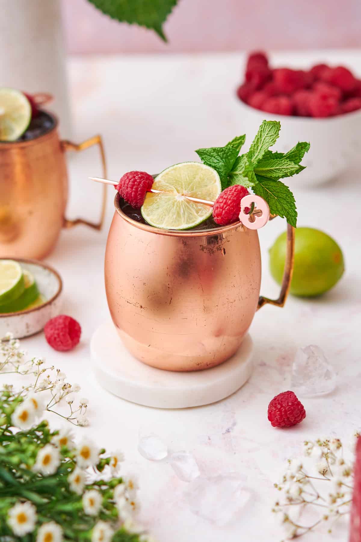 gorgeous raspberry mule cocktail in a copper mug with fresh lime wheels, raspberries, and mint surrounded by small white flowers. 