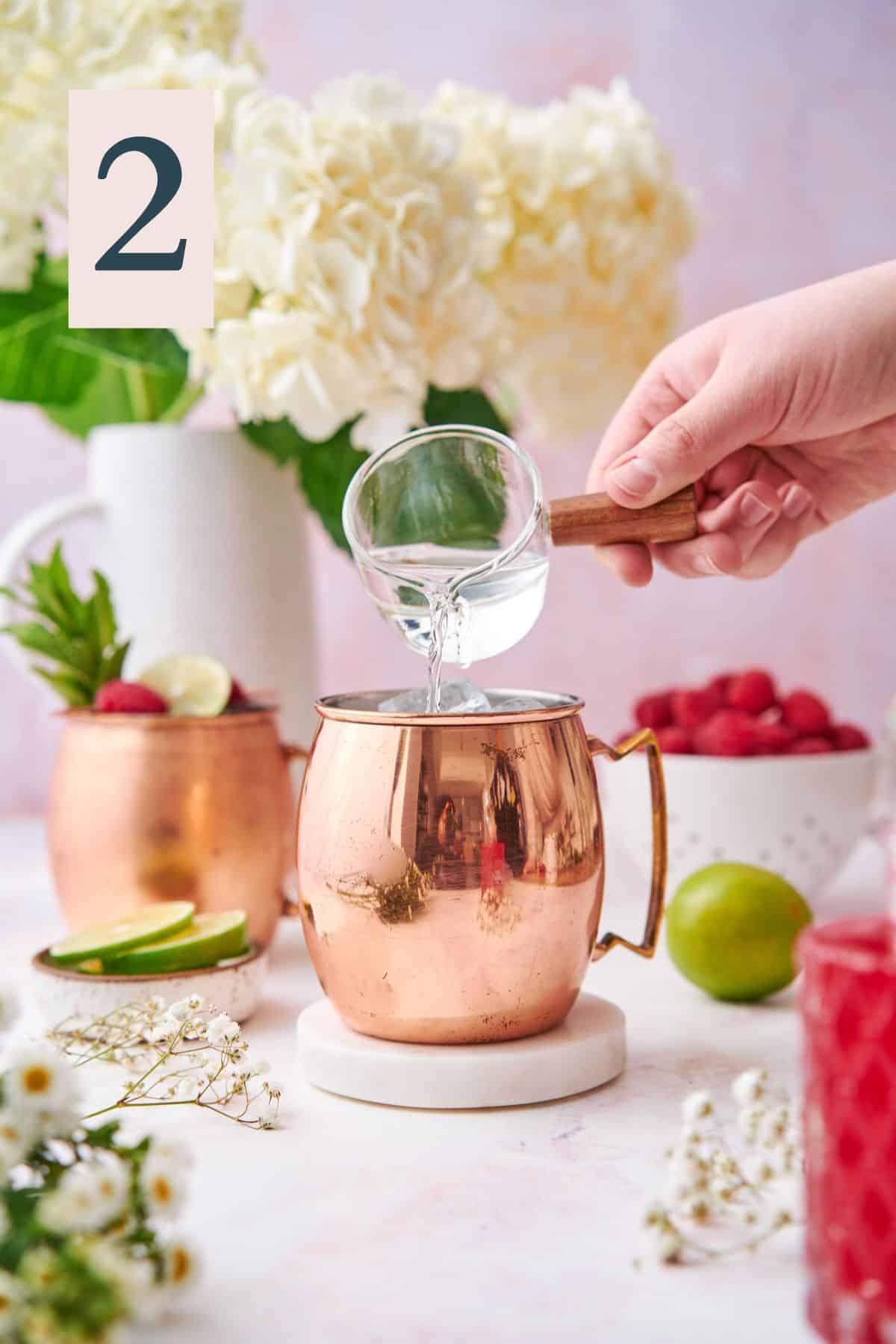 hand pouring vodka into a copper moscow mule mug with fresh raspberries, hydrangeas, and other small white flowers.  