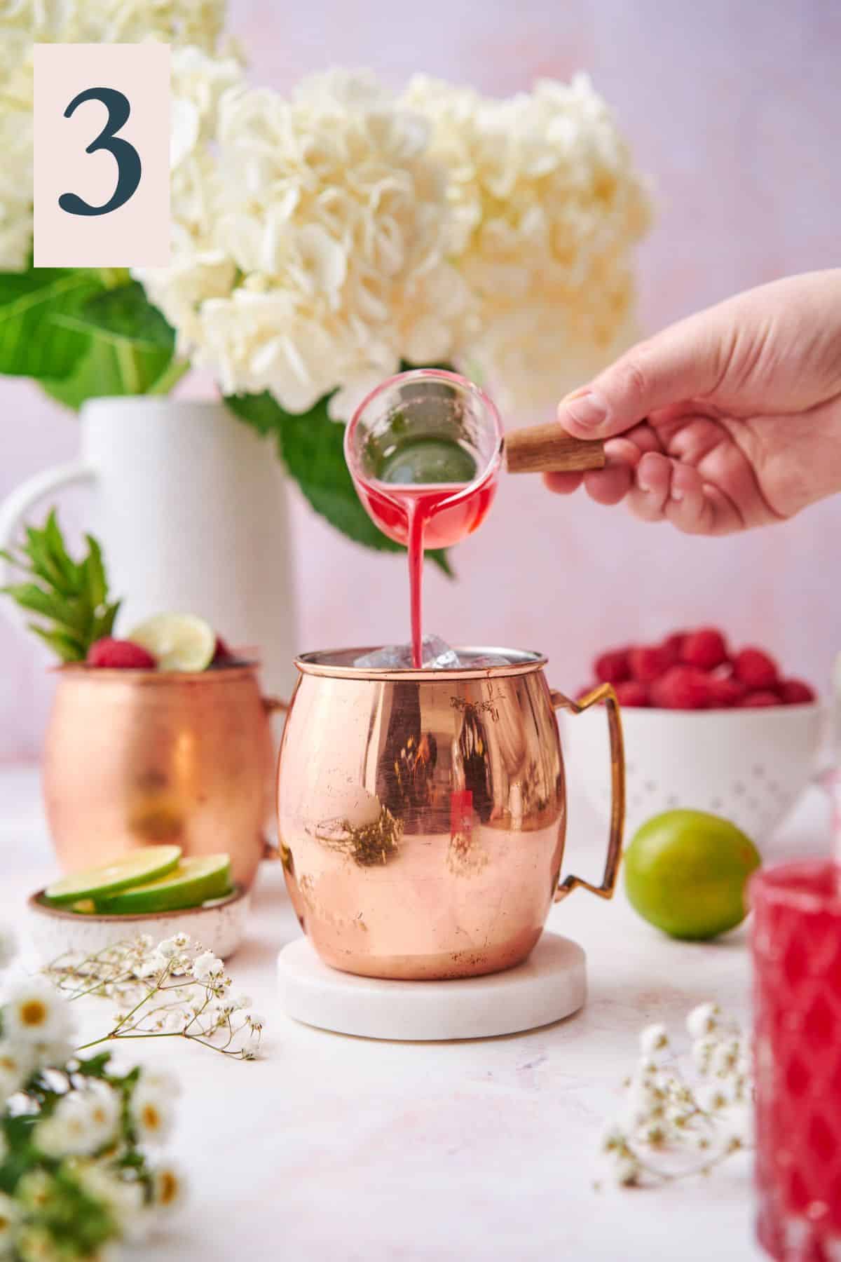 hand pouring the raspberry simple syrup into a copper moscow mule mug with fresh raspberries, hydrangeas, and other small white flowers.  