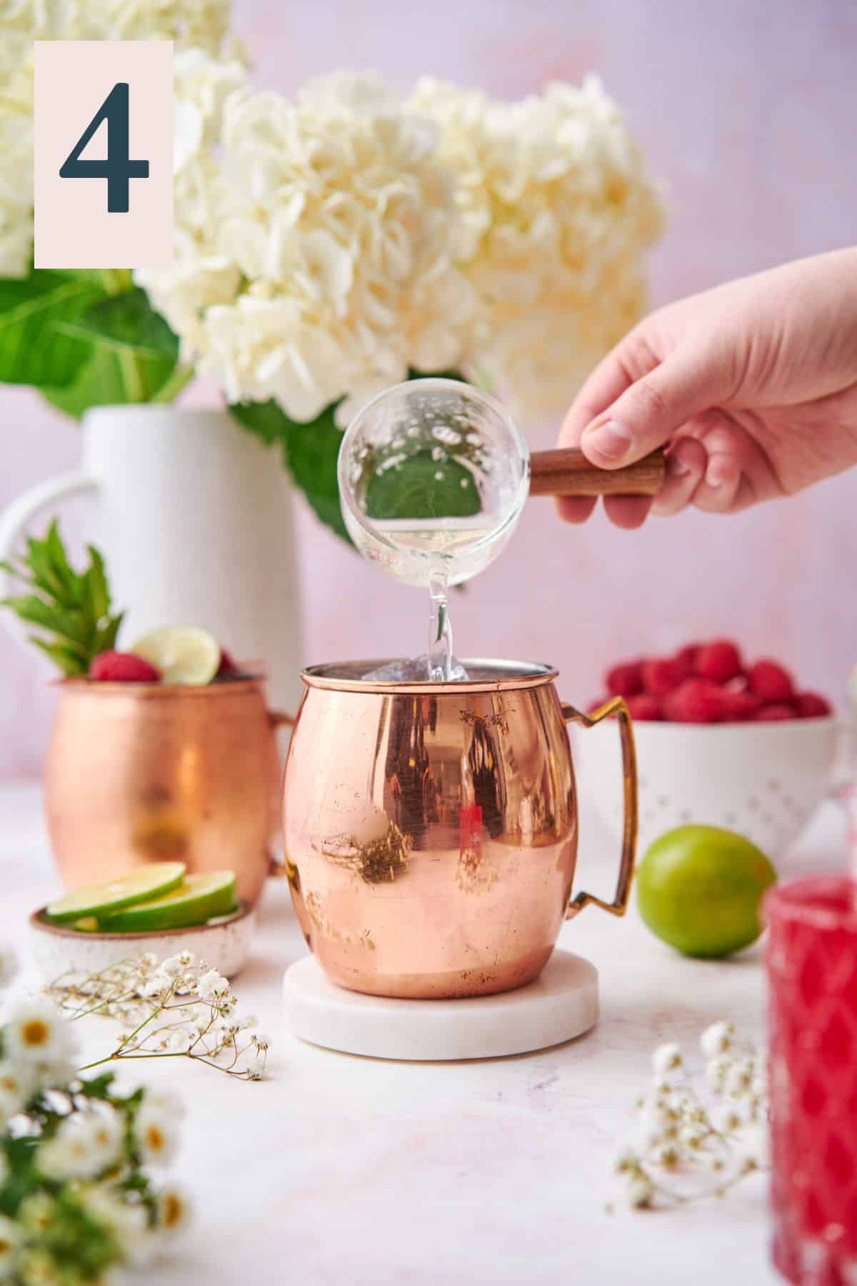 hand pouring ginger beer into a copper moscow mule mug with fresh raspberries, hydrangeas, and other small white flowers.  