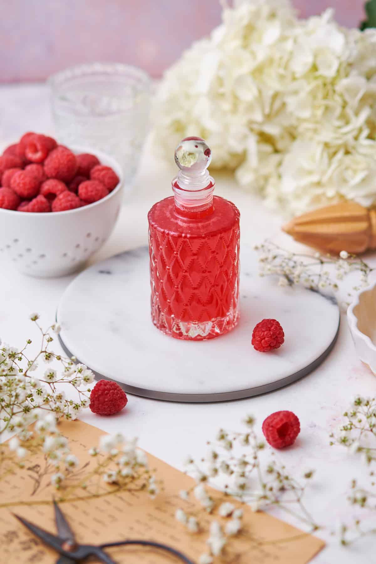 Small cute jar of raspberry simple syrup surrounded by fresh raspberries, hydrangeas, and baby's breath.  