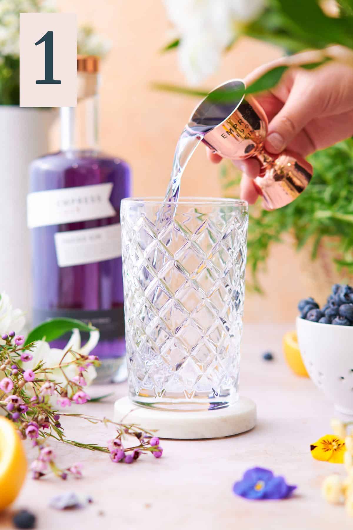 hand pouring purple Empress gin into a cocktail shaker, surrounded by flowers and Empress gin in the background. 