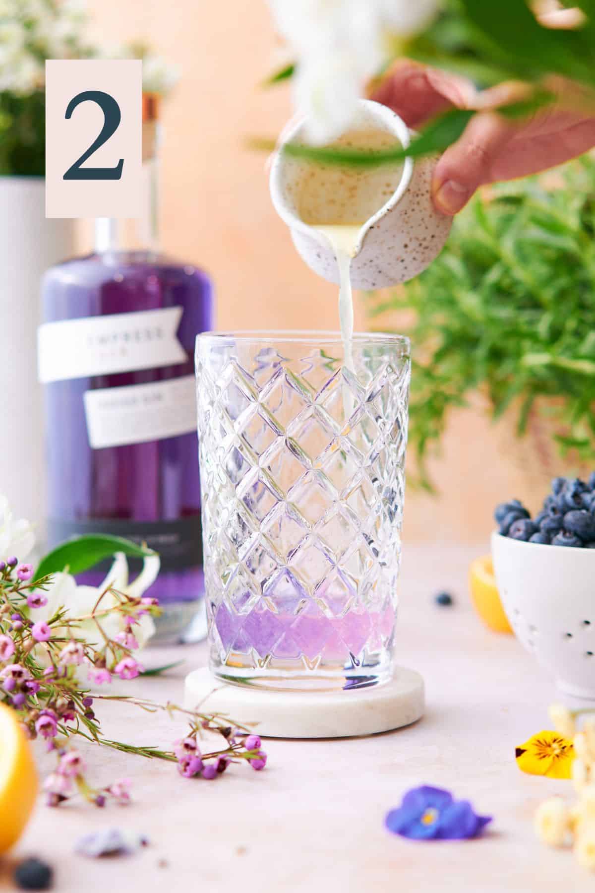 hand lemon juice into purple Empress gin in a cocktail shaker, and the gin is turning pink, surrounded by flowers and Empress gin in the background. 