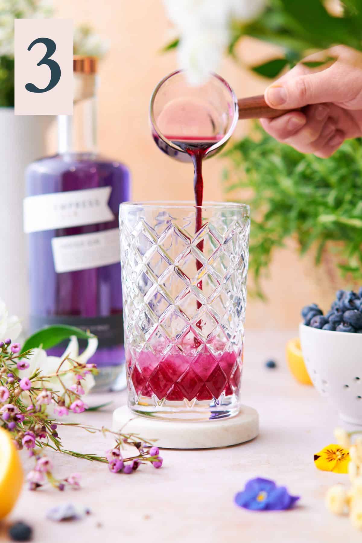 hand pouring purple blueberry simple syrup into a cocktail shaker, surrounded by flowers and Empress gin in the background. 