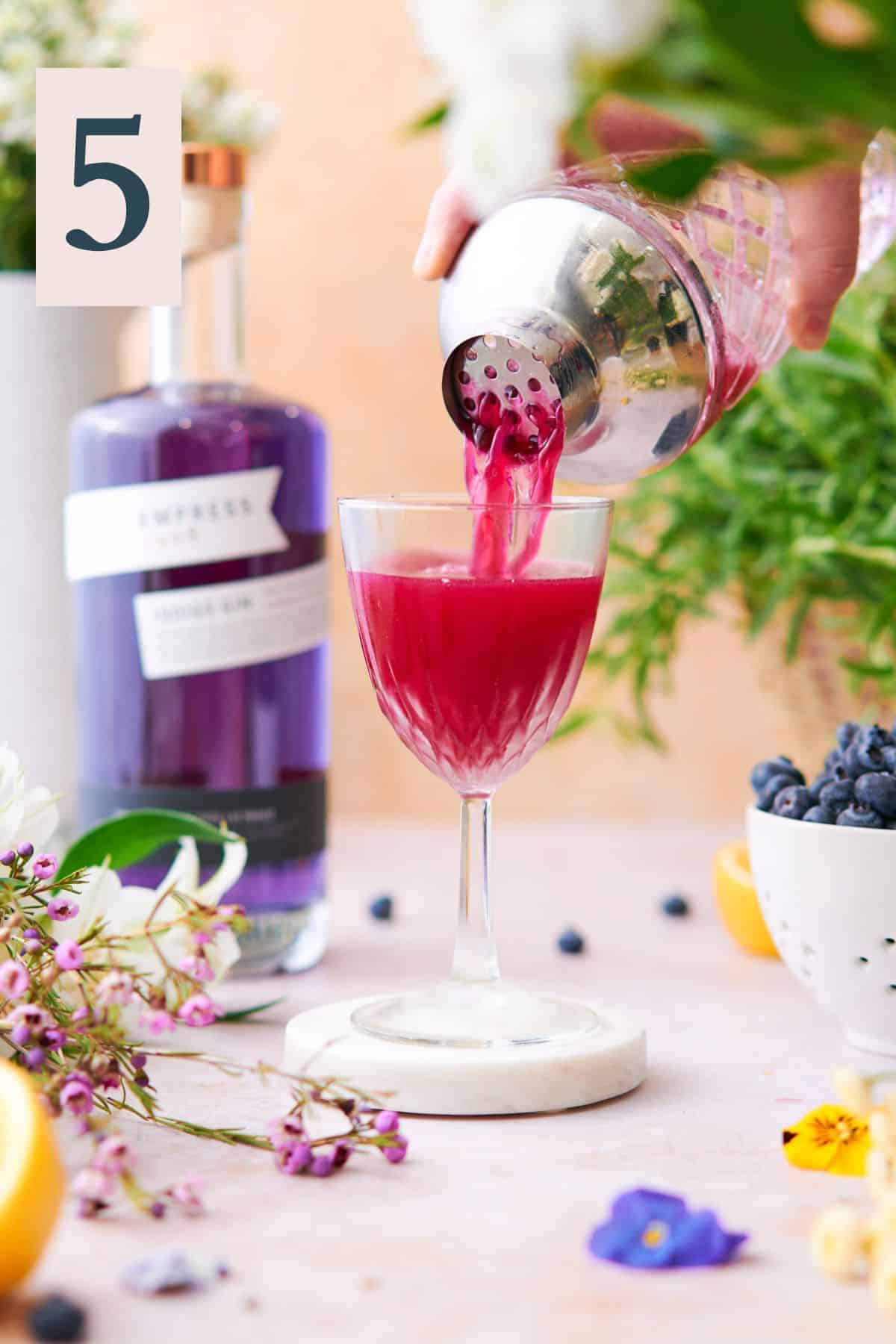 pouring a vibrant blueberry cocktail into a chilled cocktail glass, with a bottle of Empress in the background.