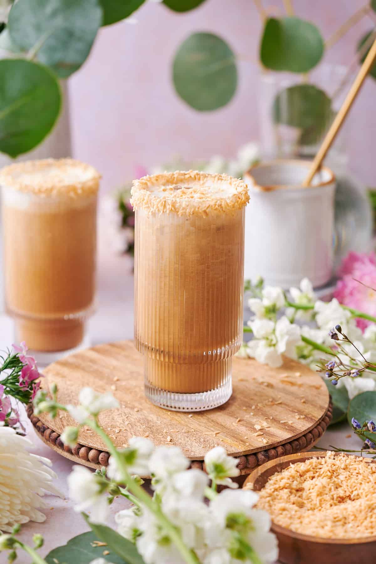 very pretty whimsical scene of coconut iced lattes surrounded by flowers and eucalyptus on a round wooden board.