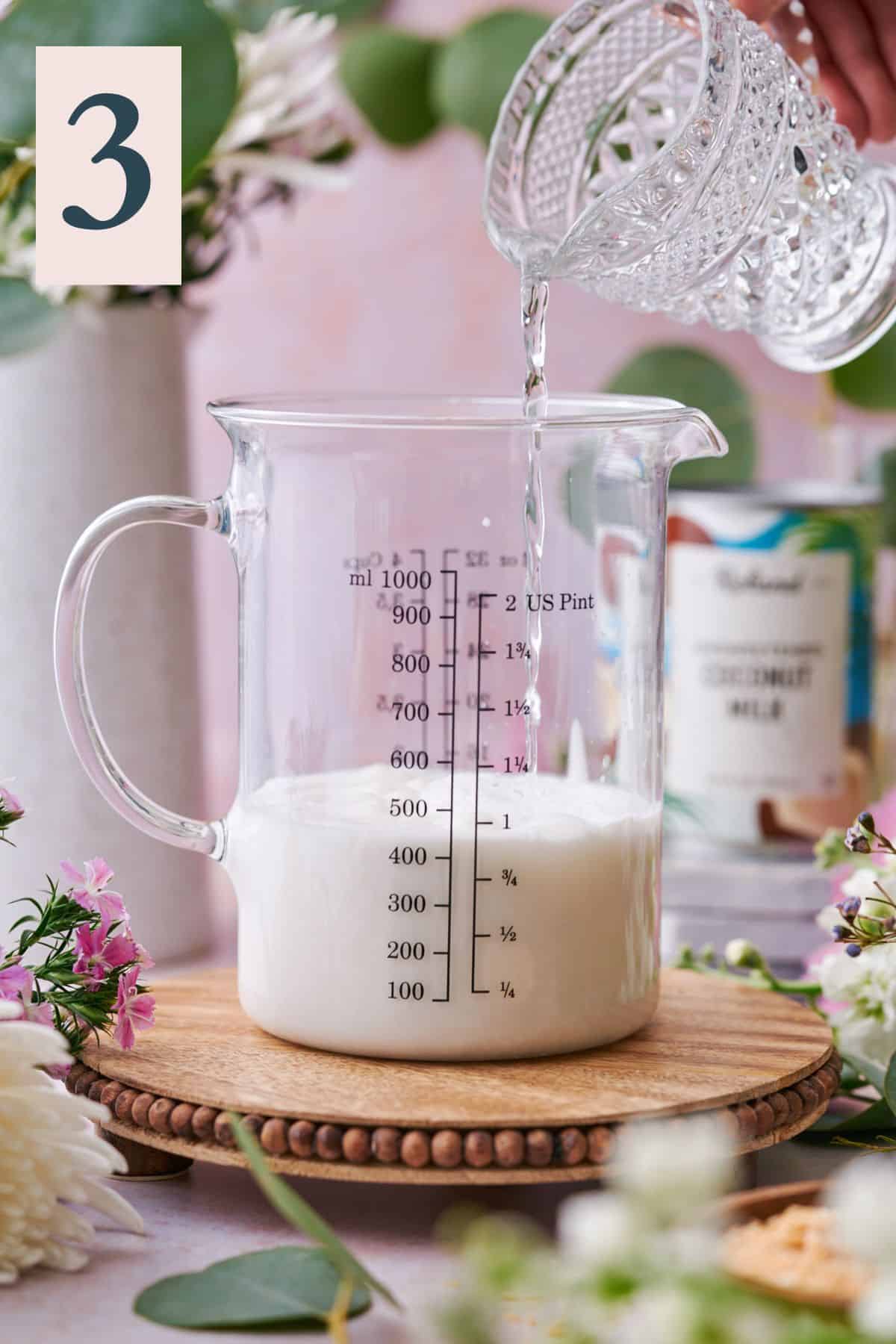 pouring water into a beaker with coconut milk, surrounded by flowers and a jar of coconut milk in the background.