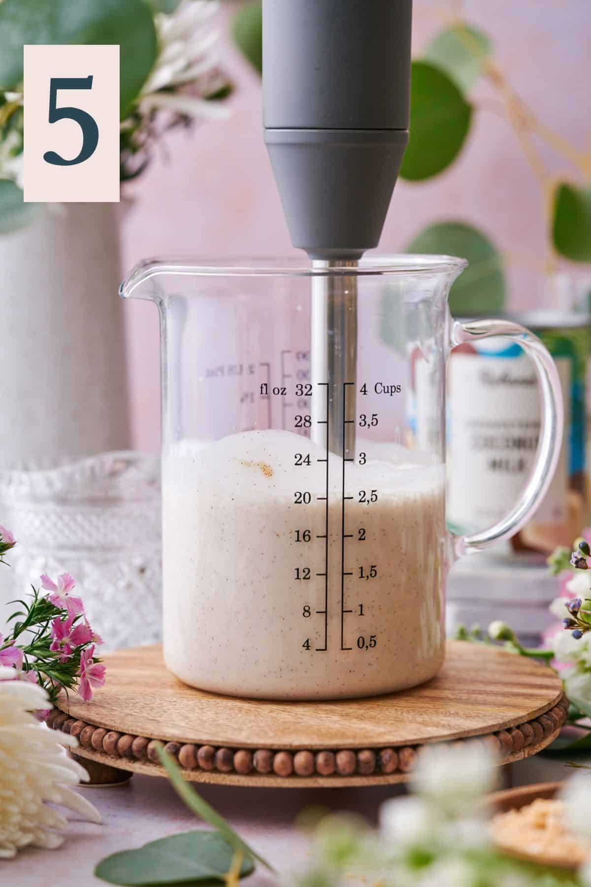 immersion blender blending up the coconut milk mixture in a large beaker surrounded by white and pink flowers and eucalyptus. 