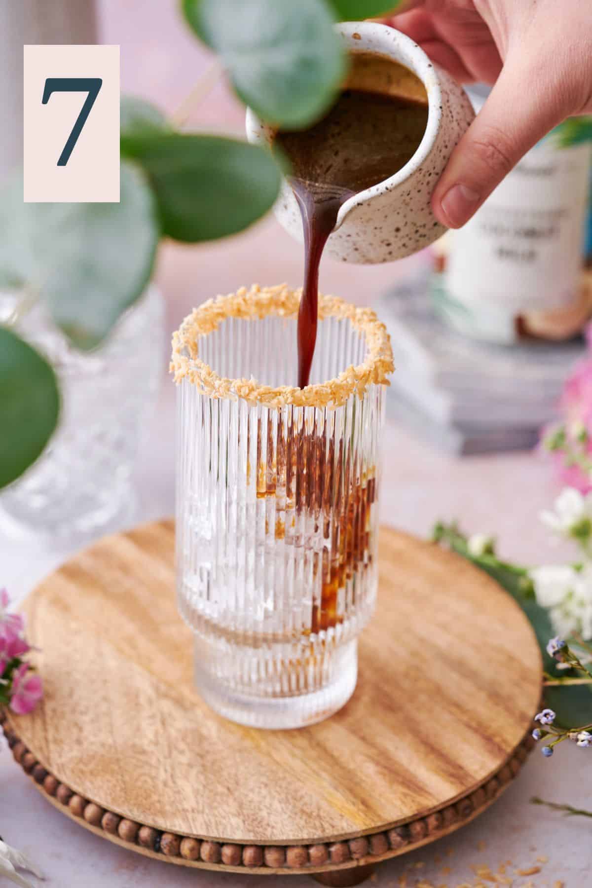 hand pouring coffee mixture into a glass with a toasted coconut rim and ice in the glass, surrounded by flowers and eucalyptus. 