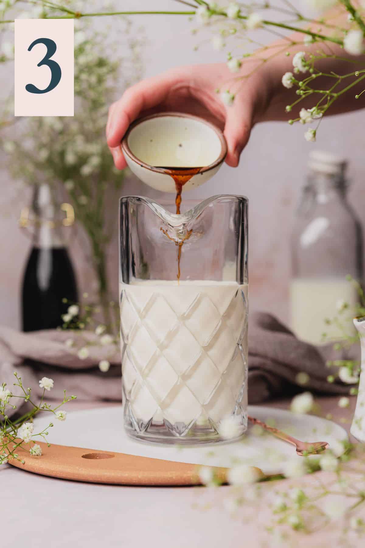 hand pouring vanilla extract into a mixing pitcher with milk and cream, surrounded by baby's breath flowers. 