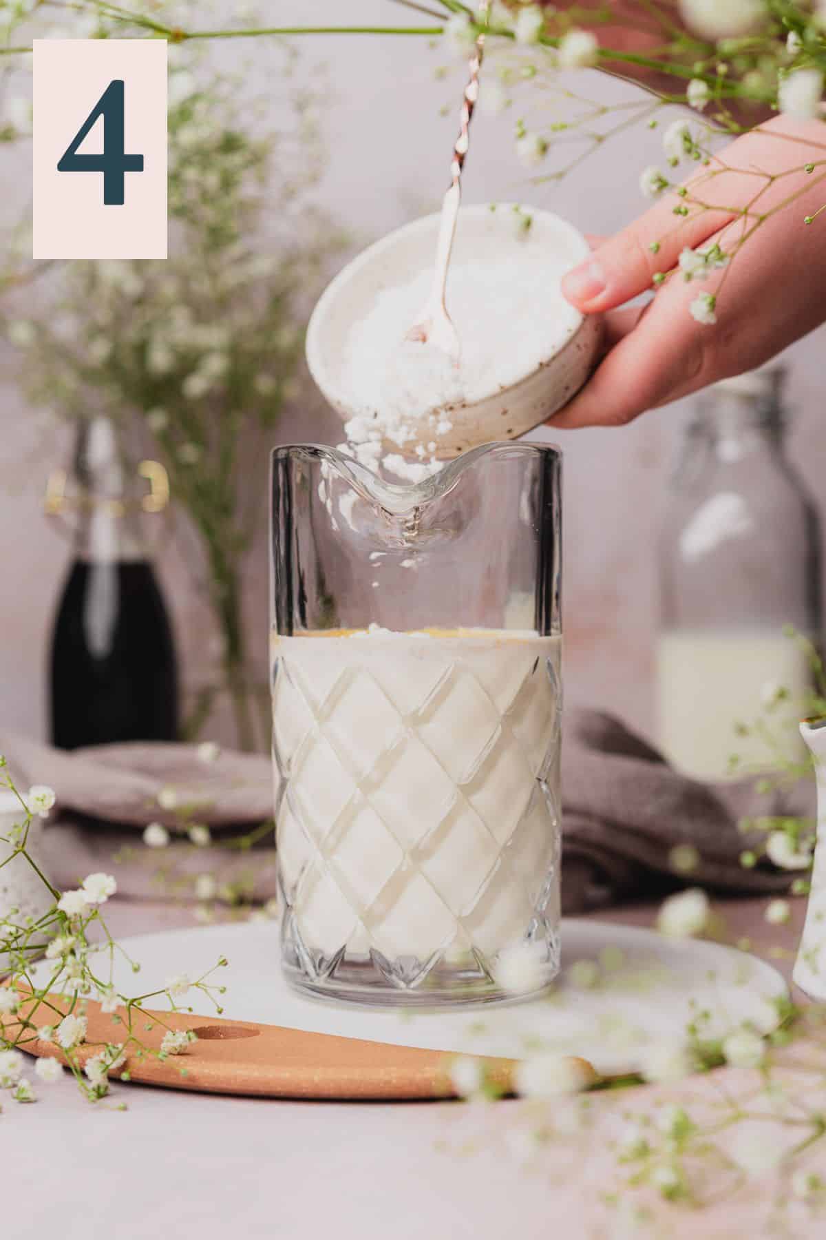 hand adding powdered sugar into a mixing pitcher with milk and cream, surrounded by baby's breath flowers. 