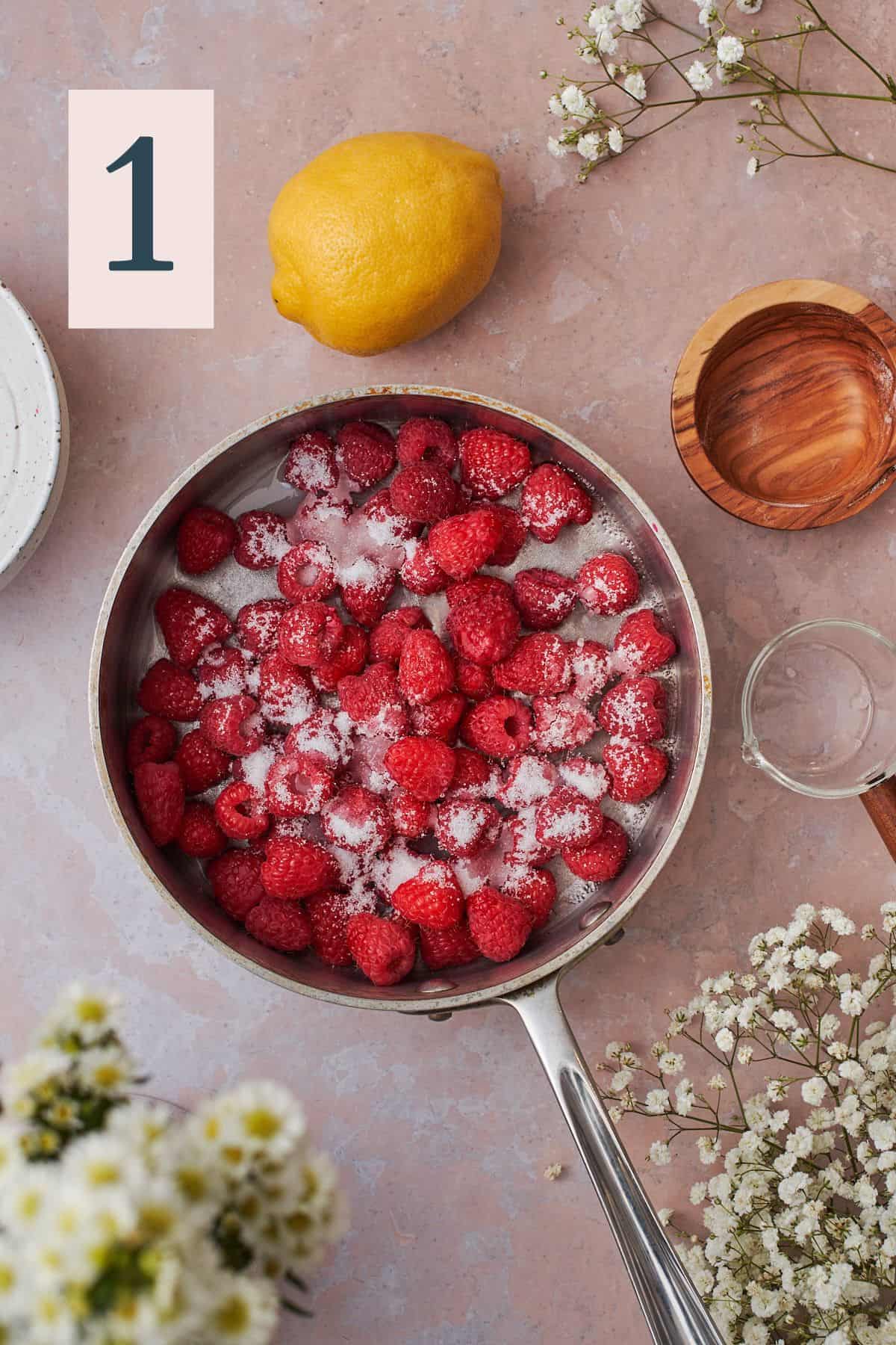 raspberries in a pan with sugar surrounded by a lemon and white flowers. 