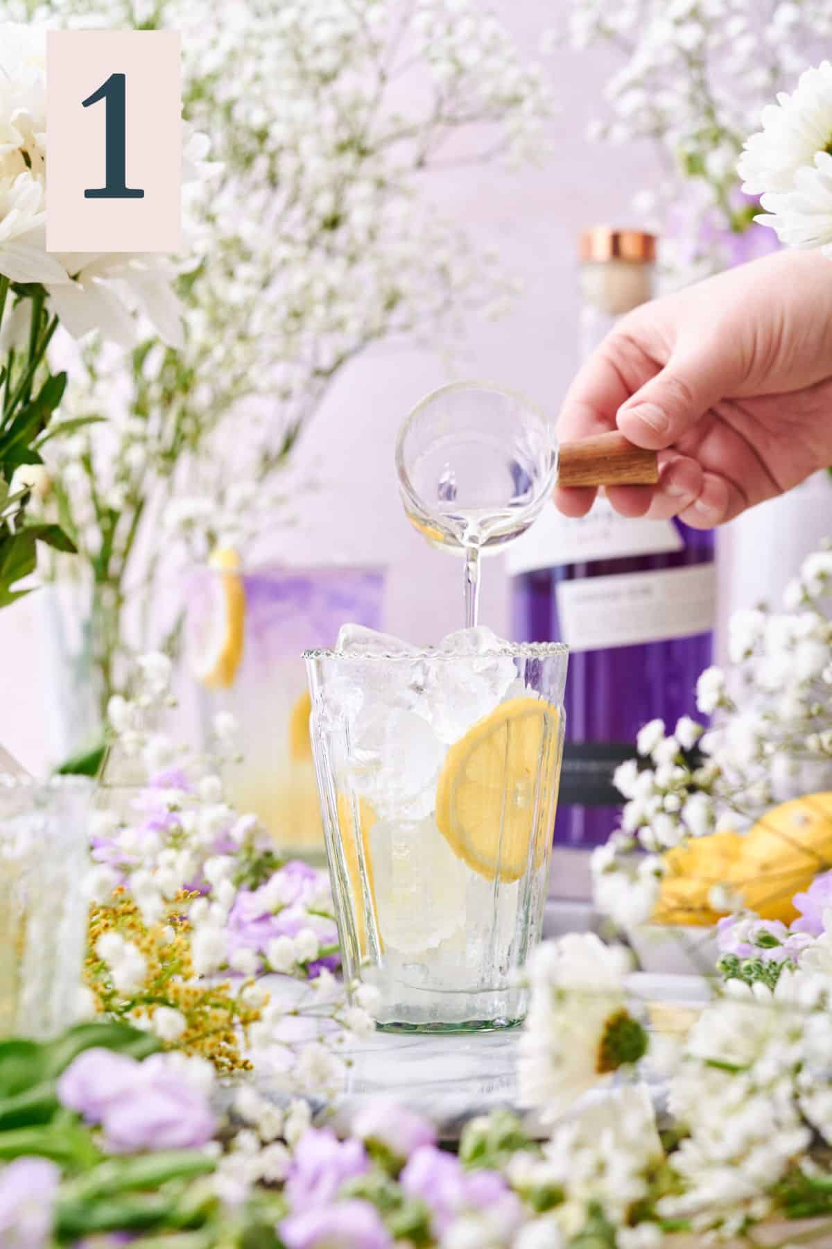 hand pouring simple syrup into a glass with lemon wheels and ice surrounded by lots of white and purple flowers.